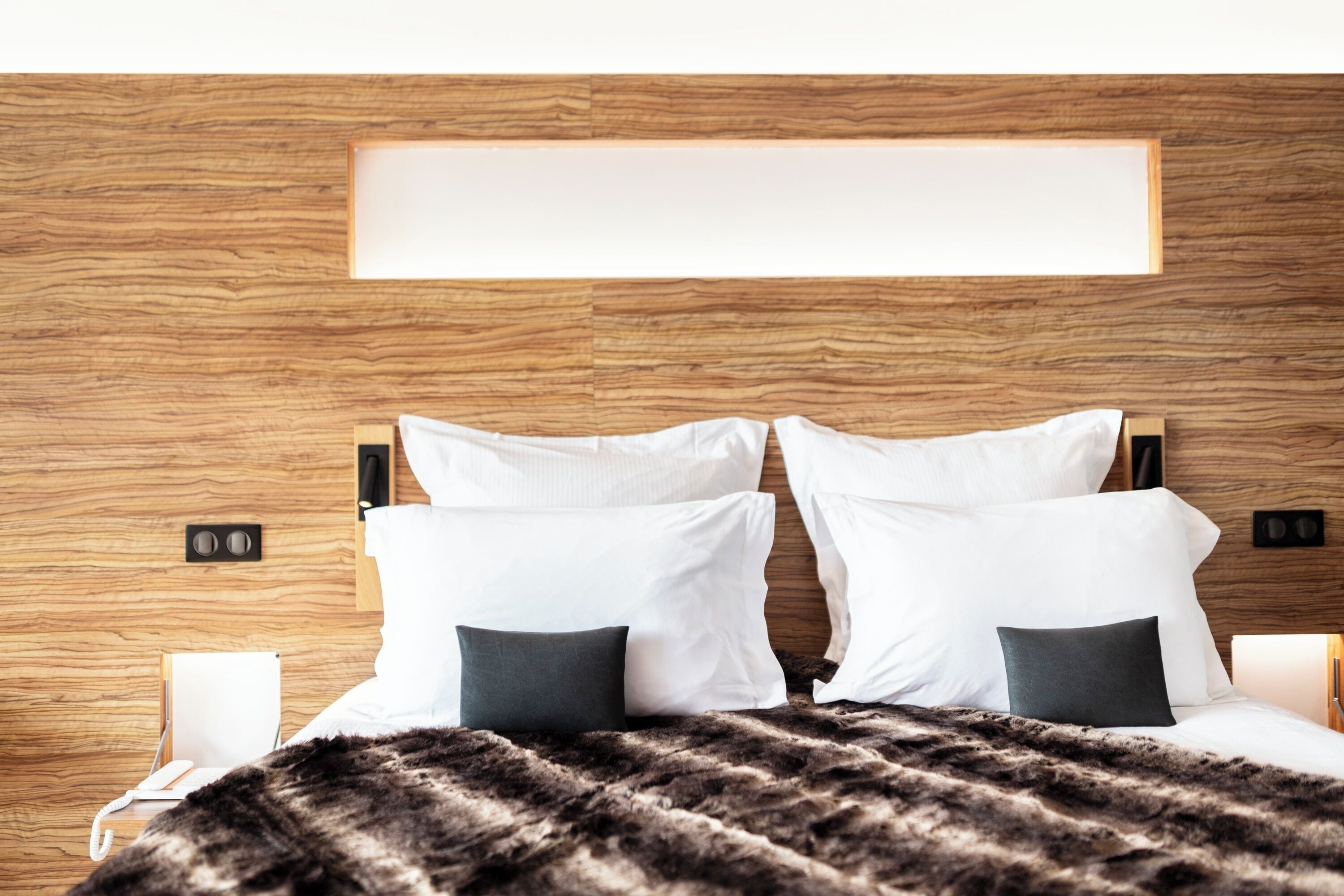Una habitación de hotel moderna con cabecera de madera, una cama grande con almohadas blancas y oscuras, un plaid de piel y lámparas de noche.