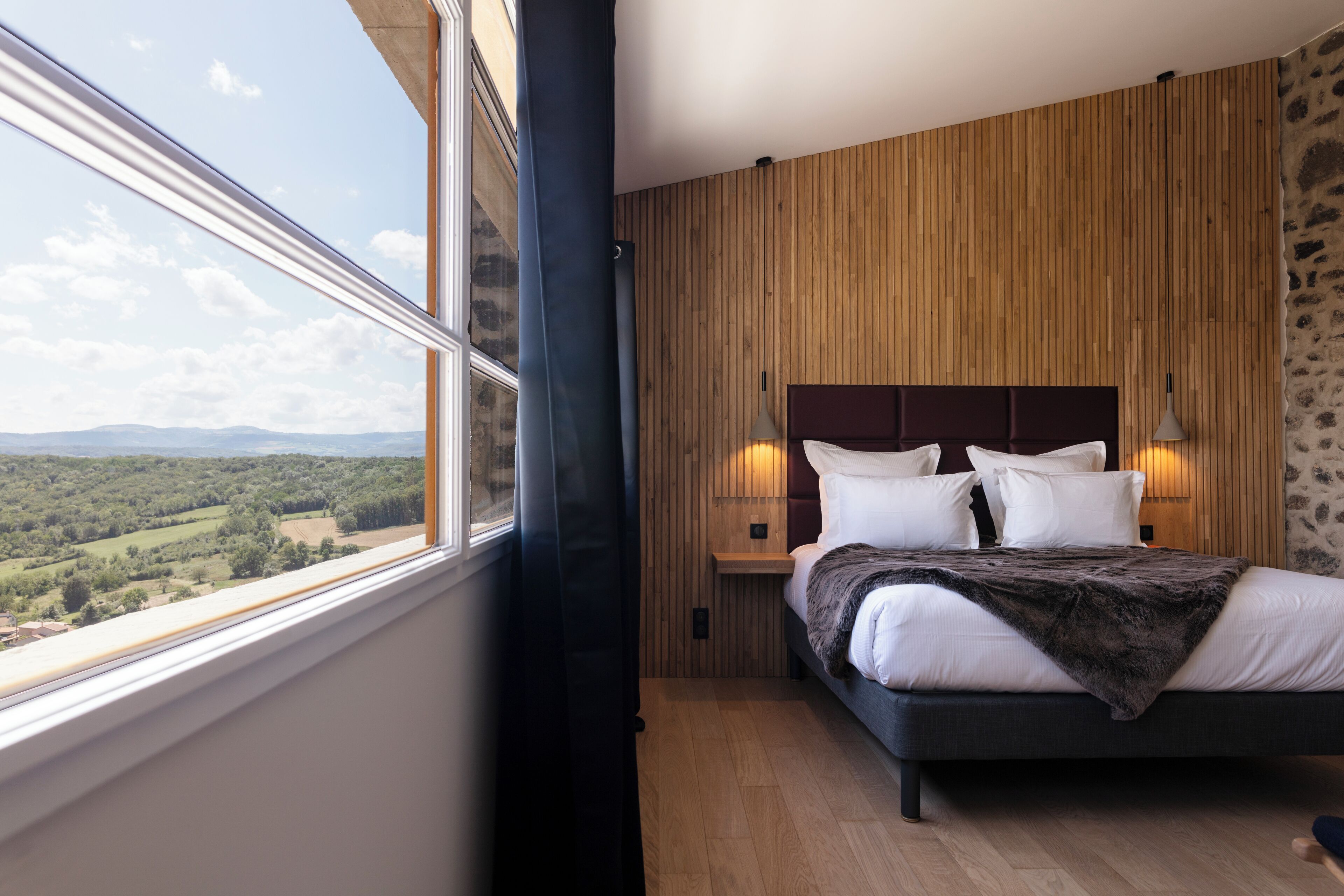 Une chambre avec une grande fenêtre, des murs lambrissés en bois, un lit avec une literie blanche et un mur en pierre.
