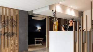 A reception area with a modern design, featuring a wooden partition with a tree design, a white reception desk, and a lamp in the background. -  Hôtel & Restaurant Origines par Adrien Descouls - Teritoria (Le Broc)