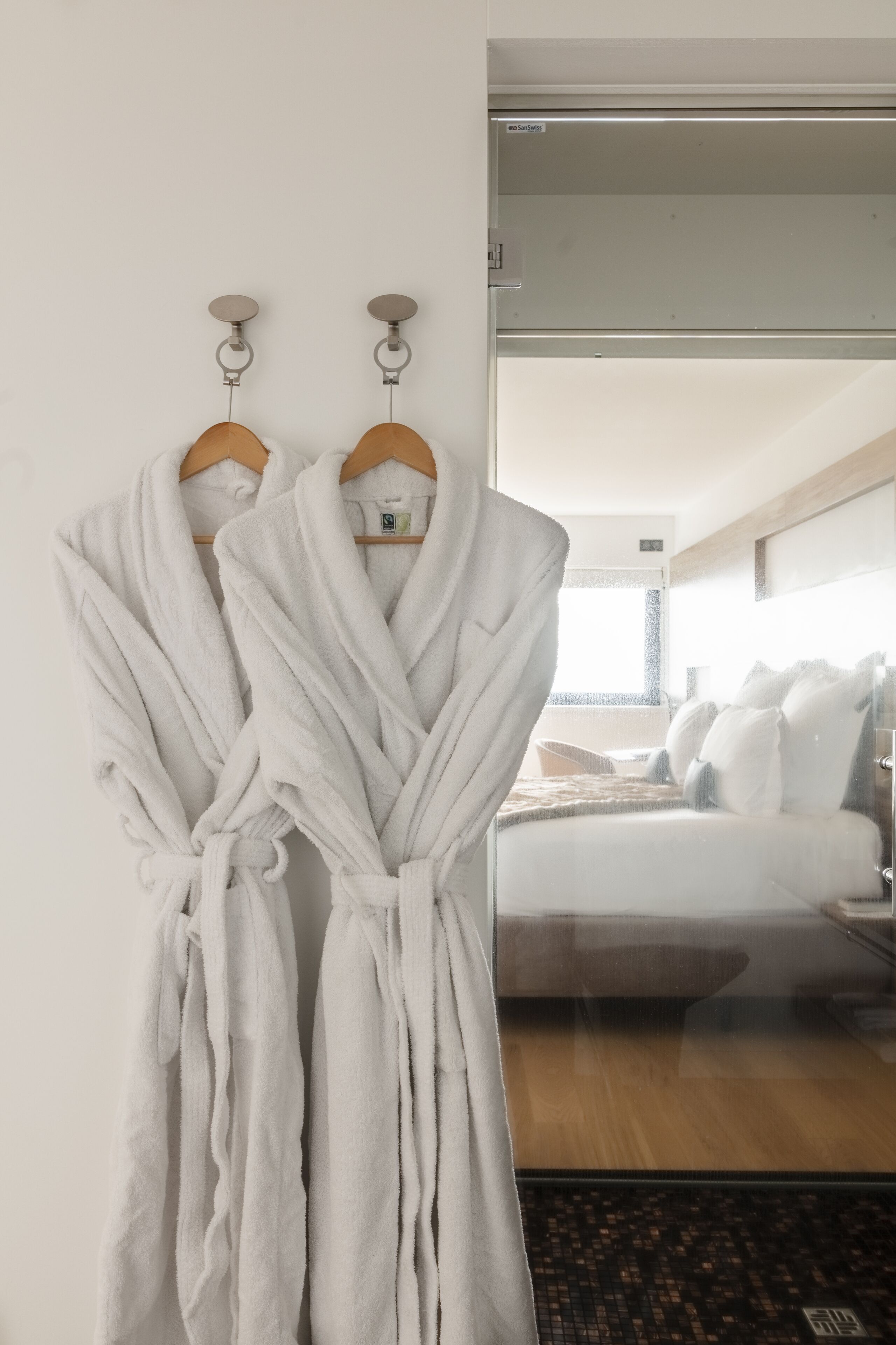 Deux peignoirs blancs sont accrochés à un mur, un lit est recouvert de draps blancs et le sol est en bois.