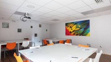 Una sala de conferencias con una mesa grande, sillas blancas y anaranjadas, y un proyector en el techo.