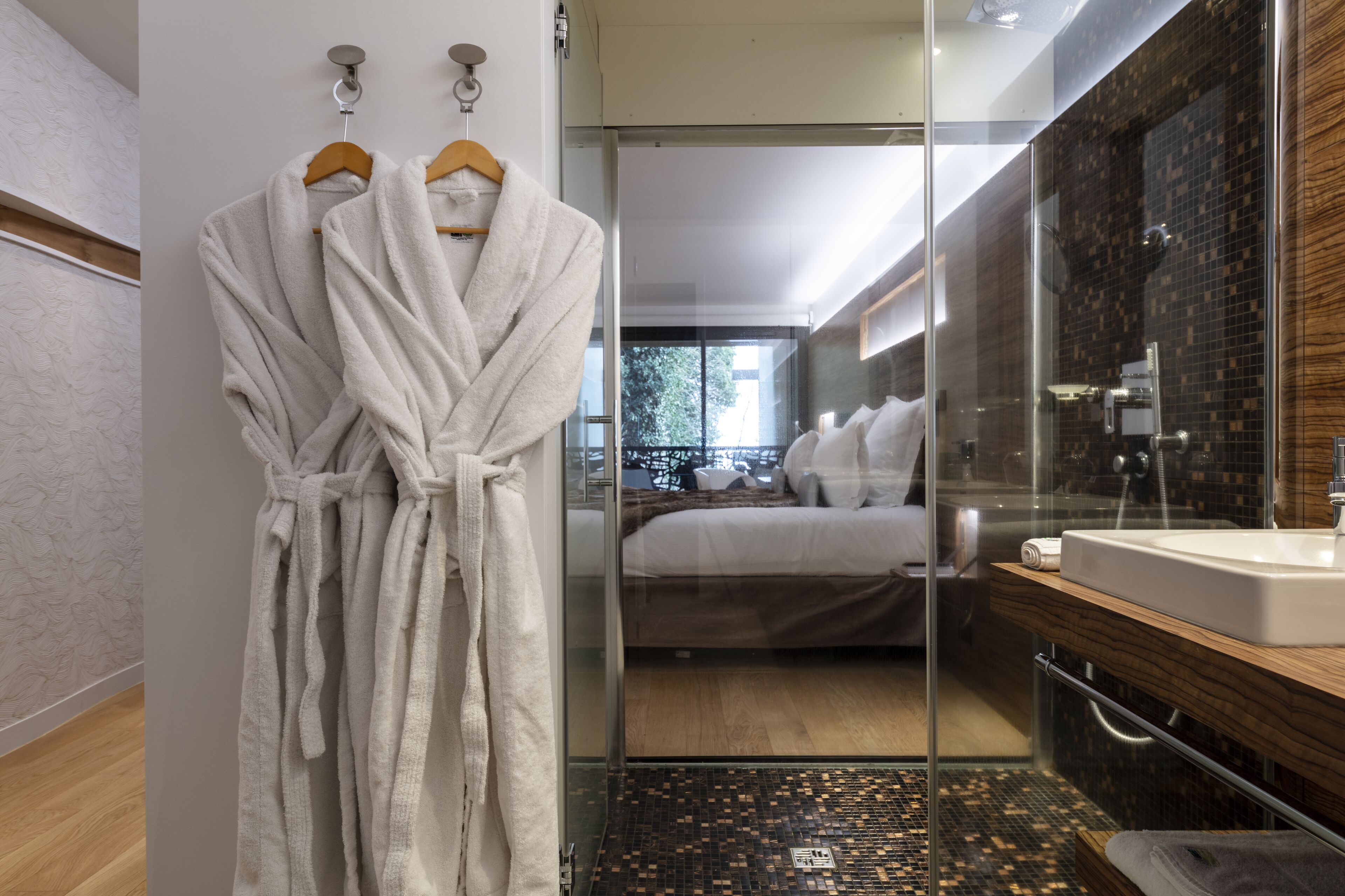 A modern bathroom with a glass shower enclosure, a wooden vanity, and a bed visible through the glass.