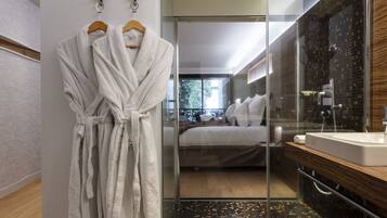 A modern bathroom with a glass shower enclosure, a wooden vanity, and a bed visible through the glass.