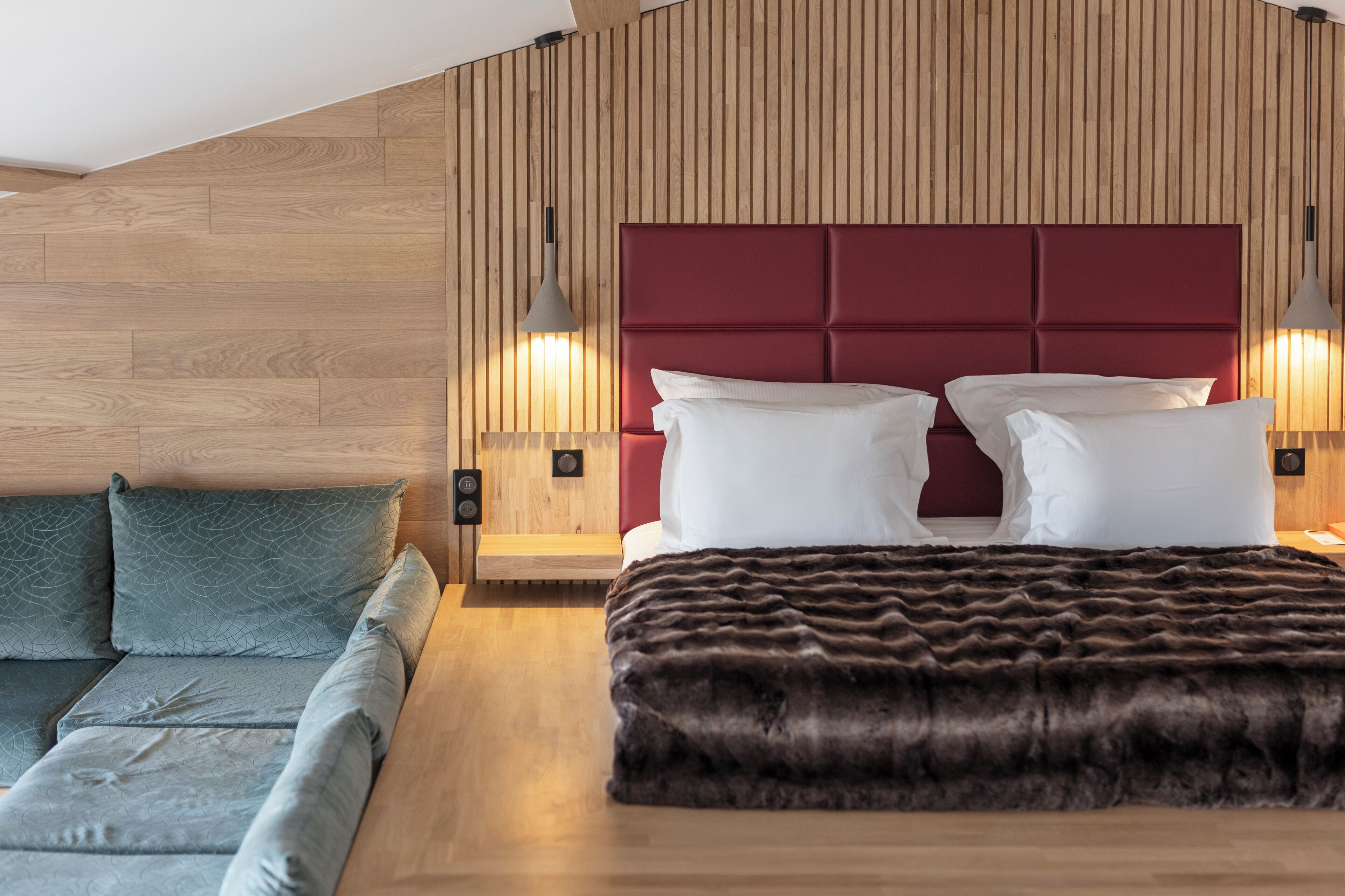 A modern bedroom with a wooden slatted wall, a red upholstered headboard, a large bed with white linens, a dark brown throw blanket, and two pendant lights.