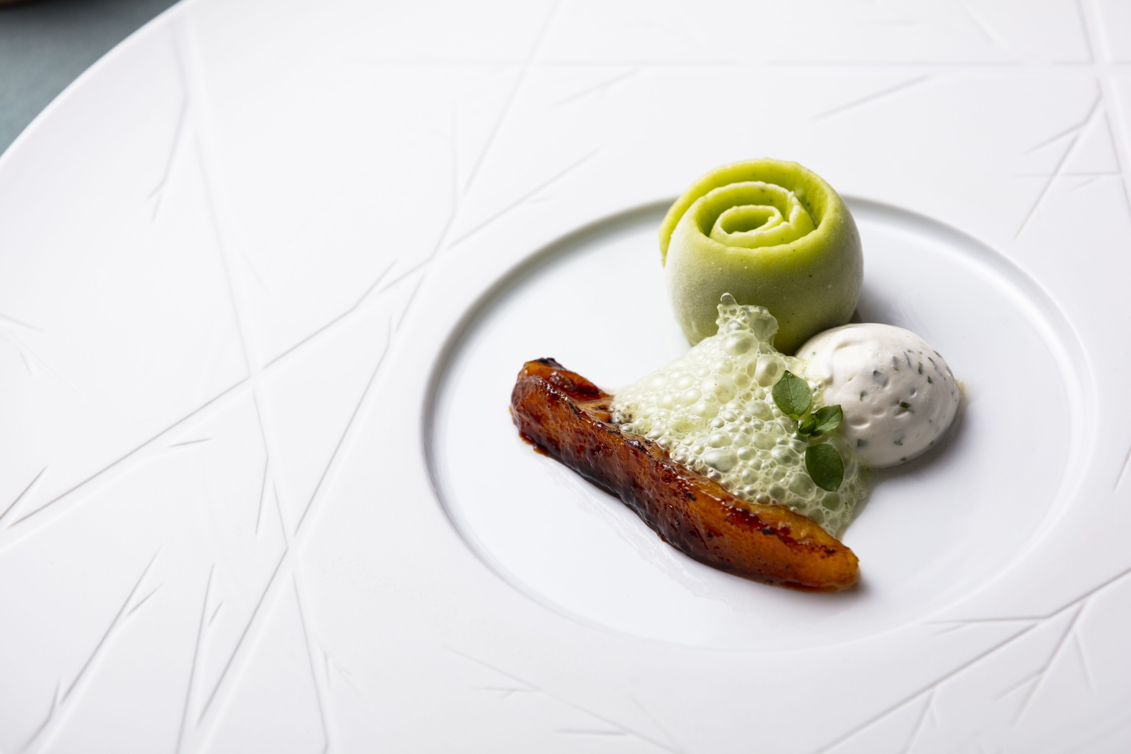 A white plate with a green rose-shaped dessert, a piece of meat, and a dollop of white sauce.