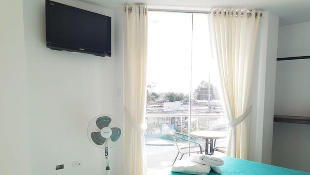 A hotel room with a bed, a standing fan, a TV mounted on the wall, and a view of the outside through a window with curtains. - Hotel Inkayacu (Paracas)