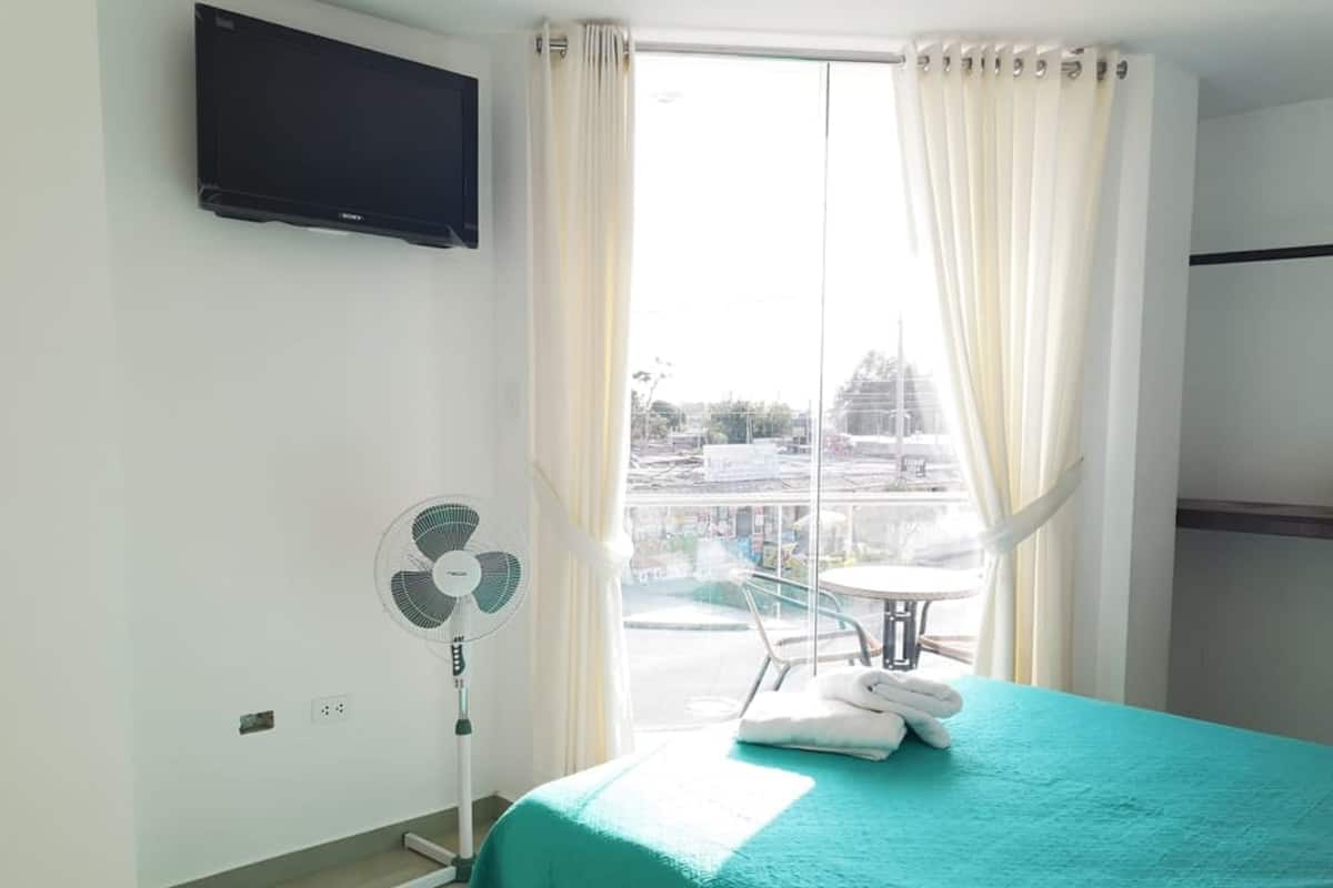 A hotel room with a bed, a standing fan, a TV mounted on the wall, and a view of the outside through a window with curtains.