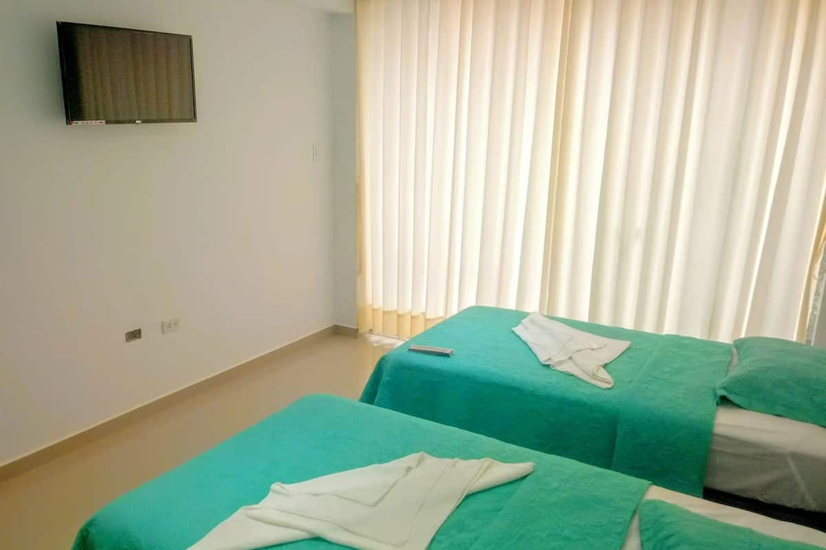 A hotel room with two single beds, each with a green bedspread and white towels. There is a flat-screen TV mounted on the wall, and a window with white curtains.