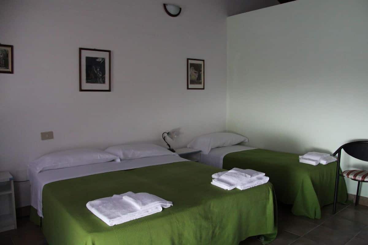 Ein Zimmer mit zwei Betten, jeweils mit grüner Bettdecke und weißen Kissen. An der Wand hängen Bilder in Rahmen, ein Sessel und eine Lampe auf einem Nachttisch.