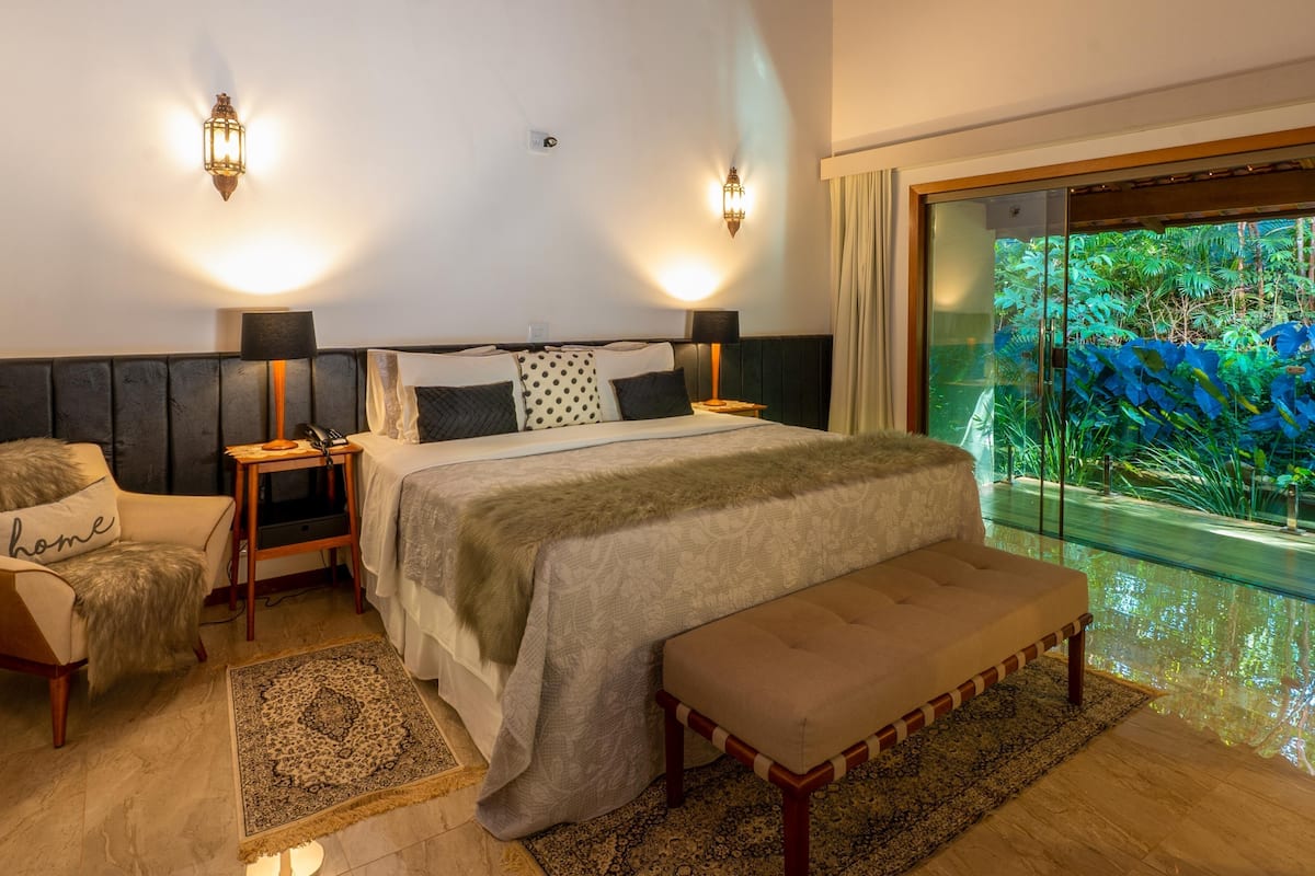 A bedroom with a bed, bedside tables, a bench, a chair, and a view of a tropical garden through a glass door.
