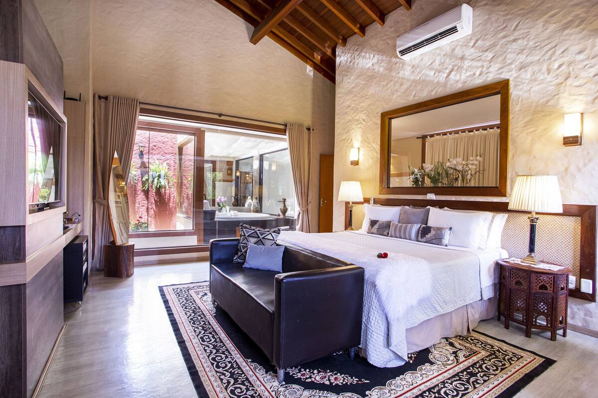 A spacious bedroom with a large bed, a sofa, and a small table. There is a large mirror above the bed and a window with curtains.