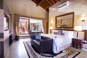 A spacious bedroom with a large bed, a sofa, and a small table. There is a large mirror above the bed and a window with curtains.