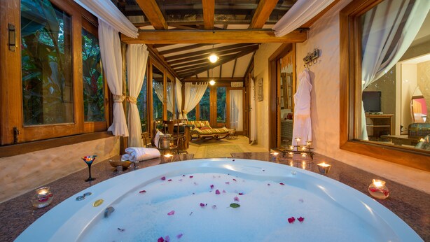 Un cuarto de spa con una bañera grande llena de agua y pétalos de rosa, vigas de madera en el techo, y vista a un dormitorio con una cama y una ventana.