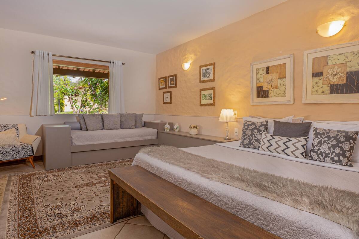 A bedroom with a bed, a sofa, a bench, a window with curtains, wall-mounted lights, and framed pictures.