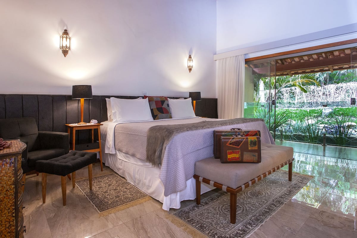 A modern hotel room with a large bed, a desk, a chair, and a view of a garden through a glass door.