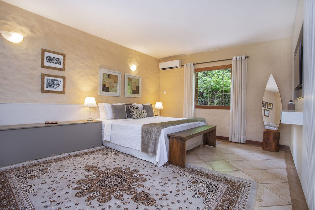 A bedroom with a bed, bedside lamps, a bench, a large rug, and framed pictures on the wall.