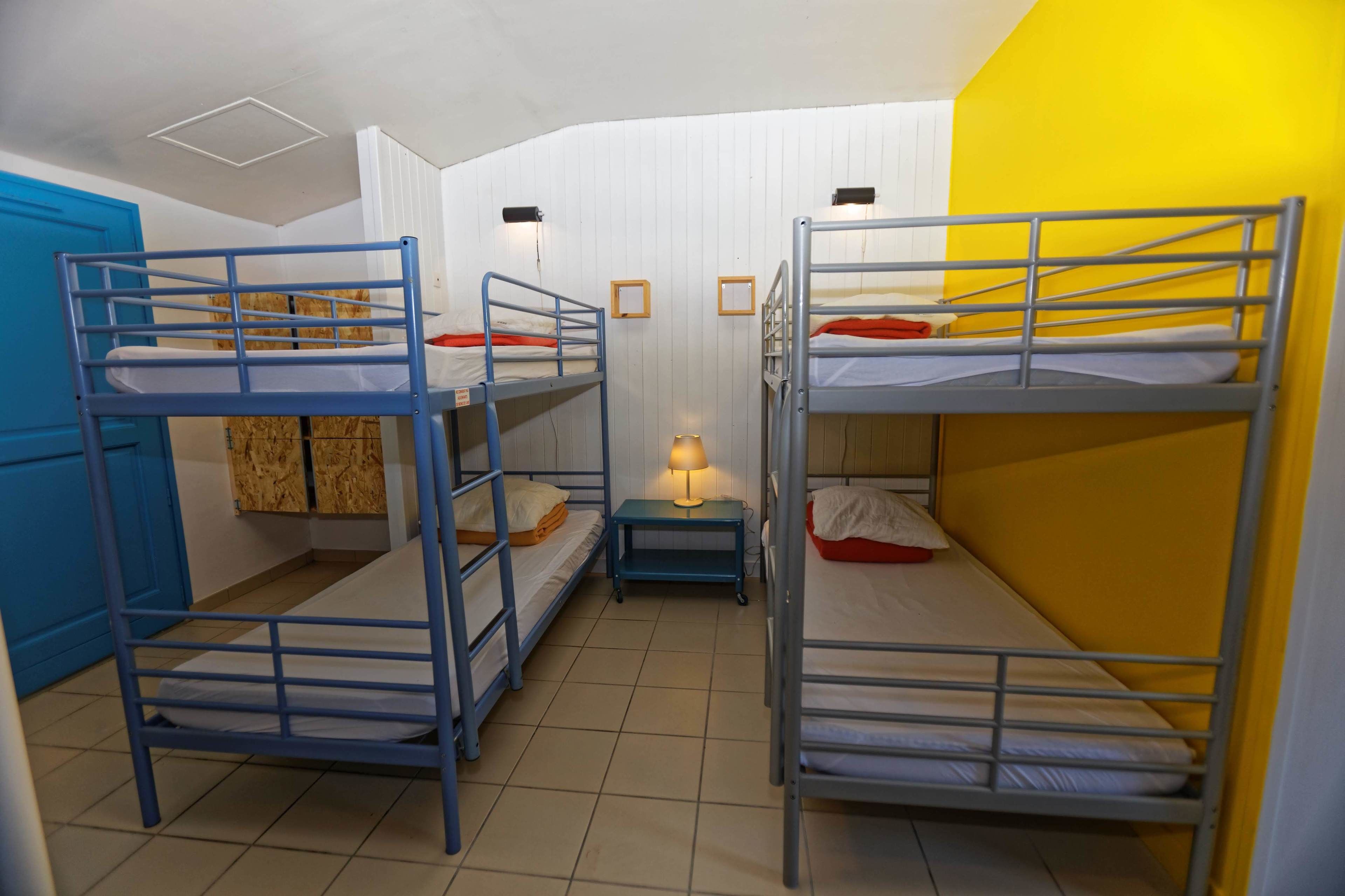 A dormitory room with bunk beds, a small table with a lamp, and a blue cabinet.