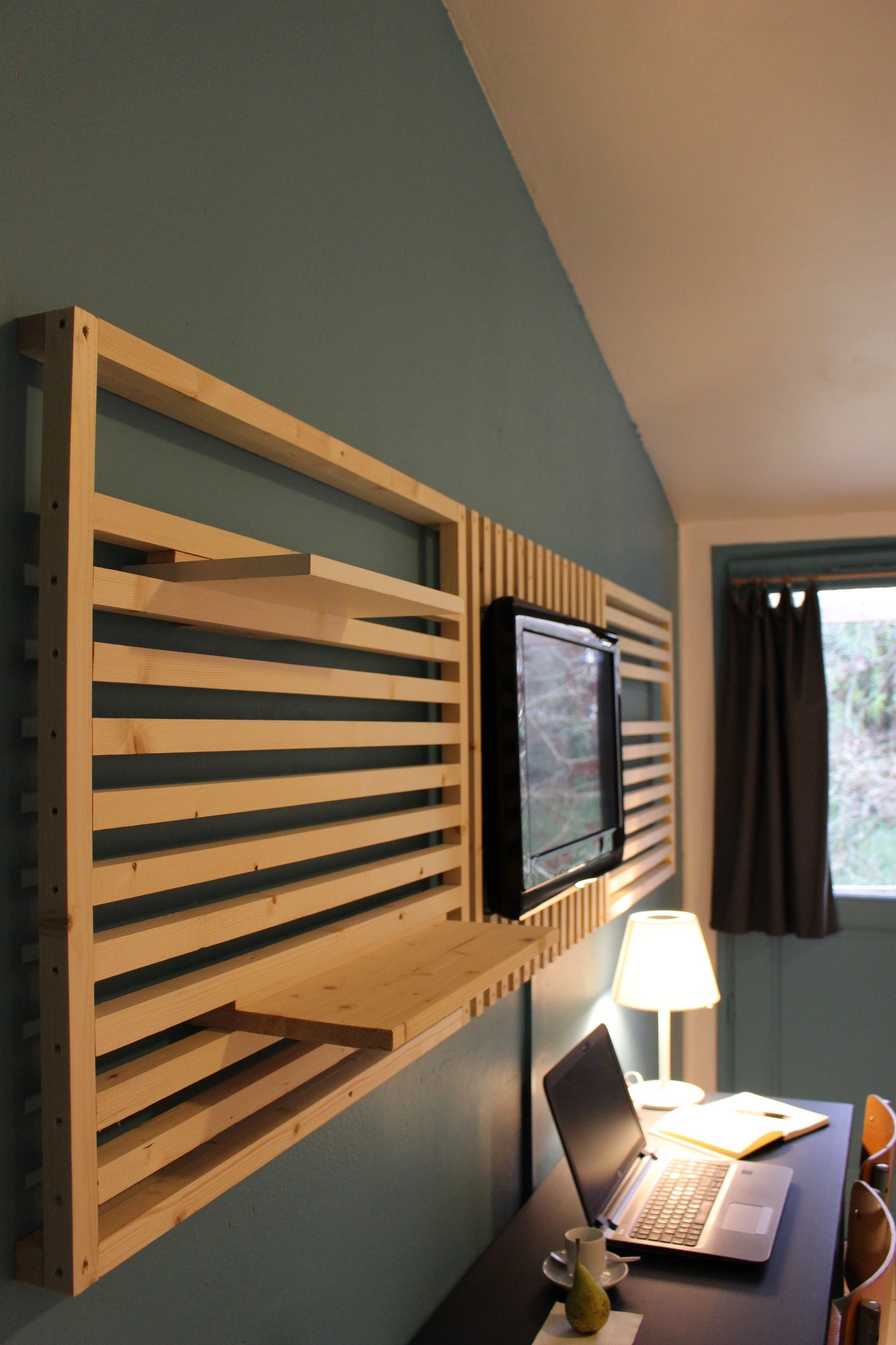 A room with a wooden slat wall-mounted TV, a desk with a laptop, a lamp, and a vase.