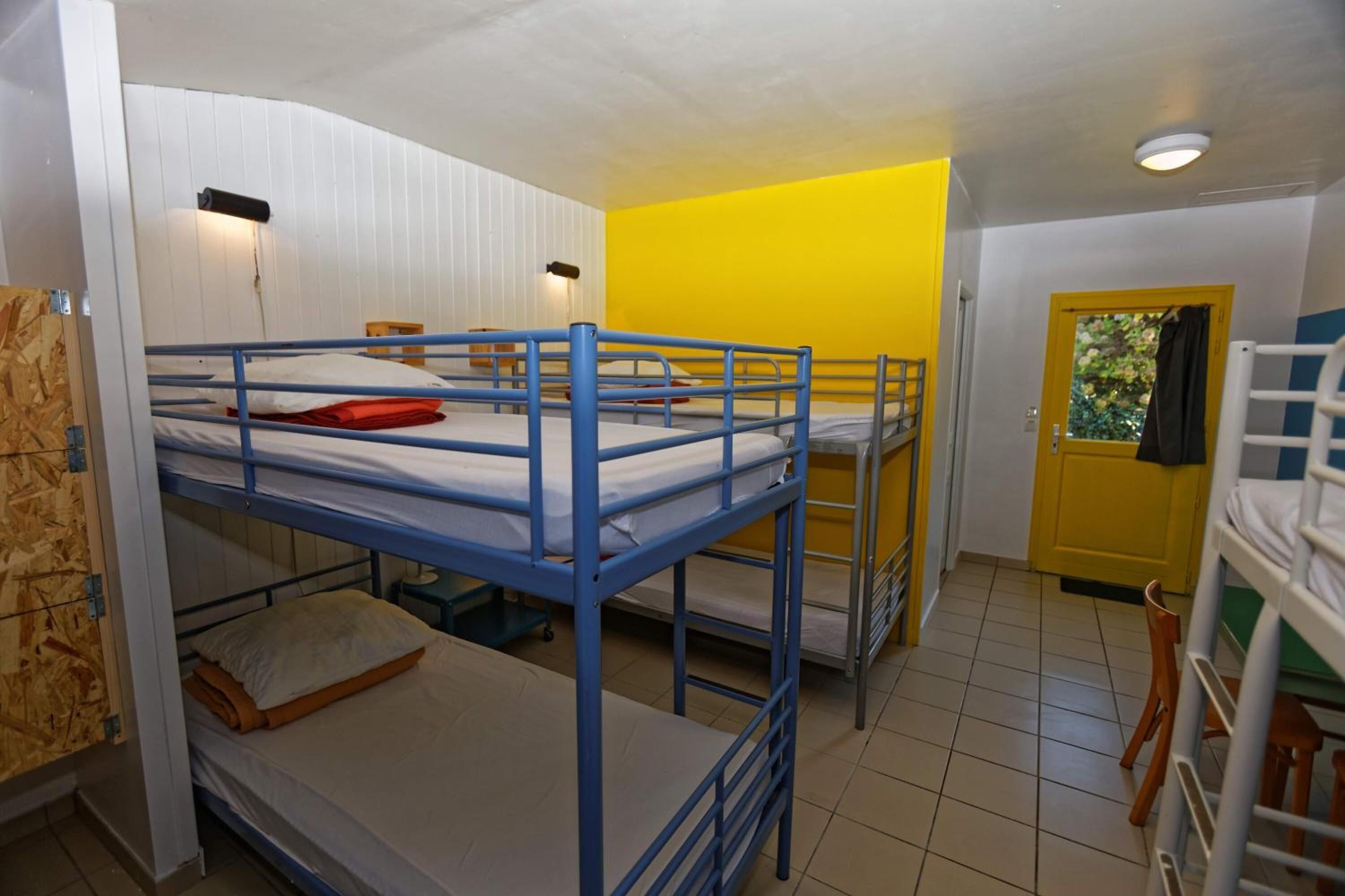 Une chambre de dortoir avec des lits superposés, un mur jaune, une porte avec une fenêtre et une petite table.