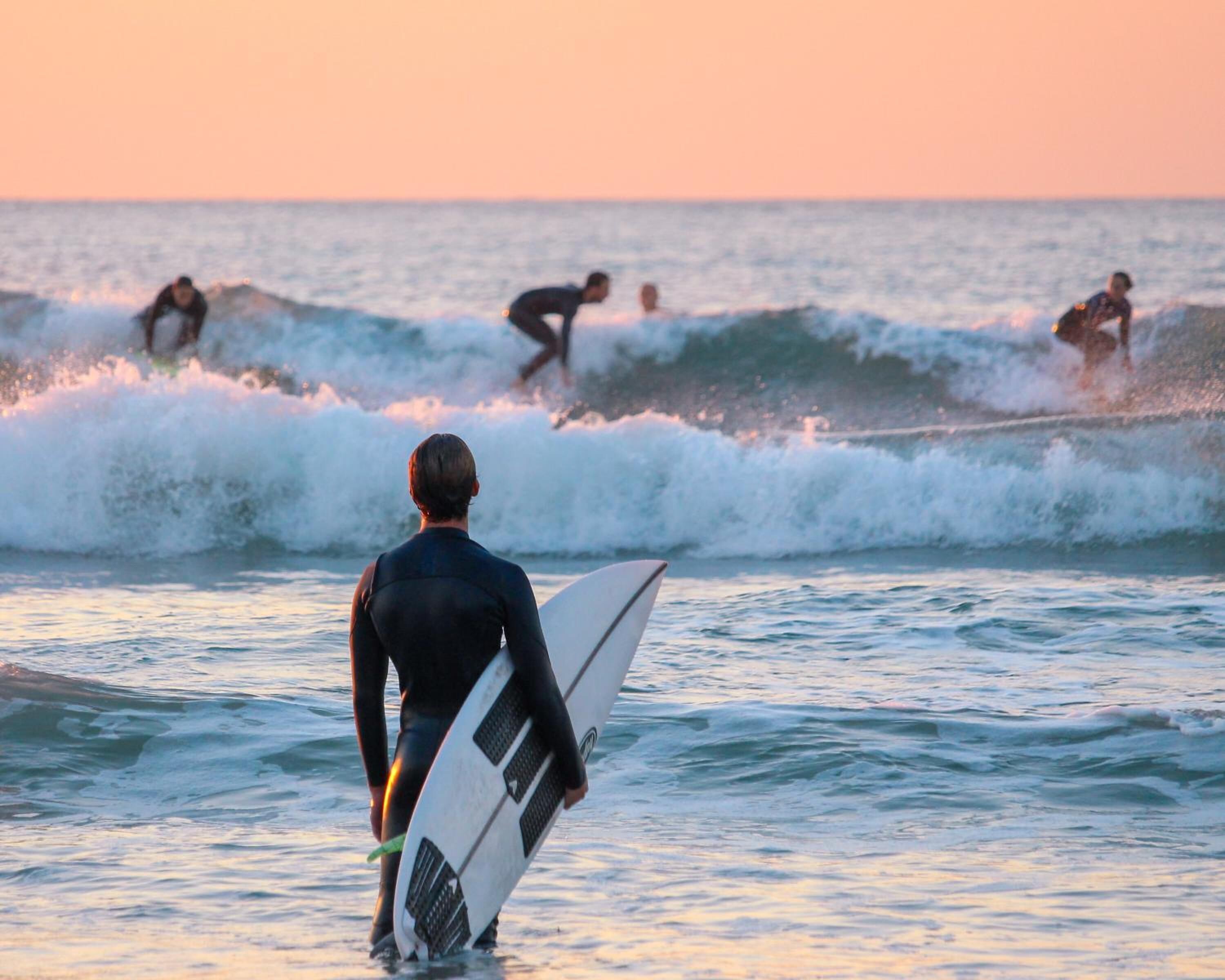 Eine Person im Neoprenanzug trägt ein Surfbrett und geht auf den Ozean zu, im Hintergrund sind Wellen zu sehen.