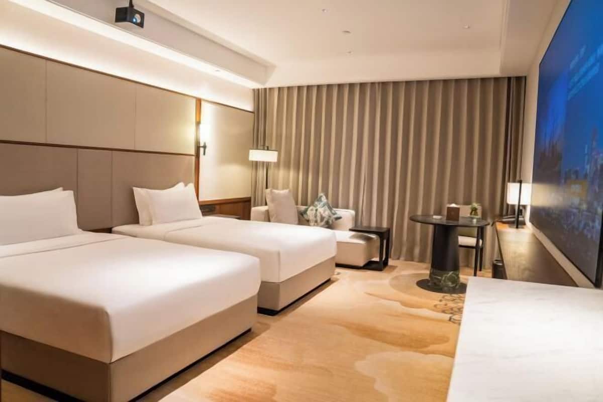 A hotel room with two beds, a small table, a lamp, and a large window with curtains.