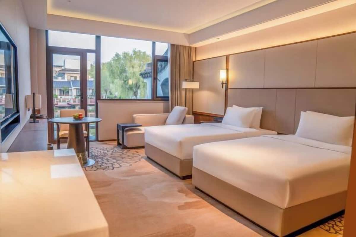 A hotel room with two beds, a small table, and a view of buildings outside.