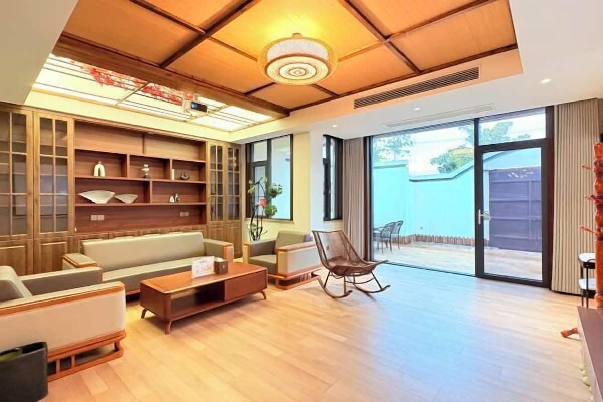 A modern living room with a sofa, coffee table, and a rocking chair. There is a large window and a door leading to an outdoor area.