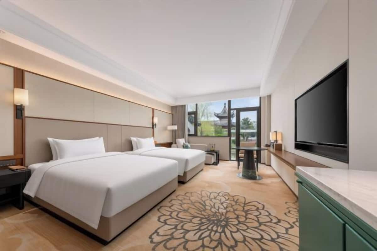 A hotel room with two beds, a TV, a desk, and a view of greenery outside.
