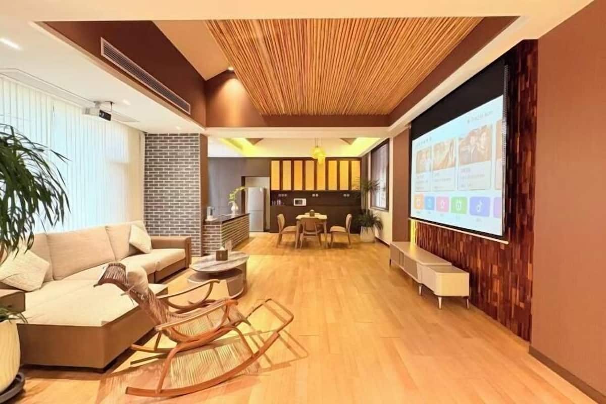 A modern living room with a sofa, a coffee table, a wooden rocking chair, a dining area with a table and chairs, and a large screen displaying a presentation.