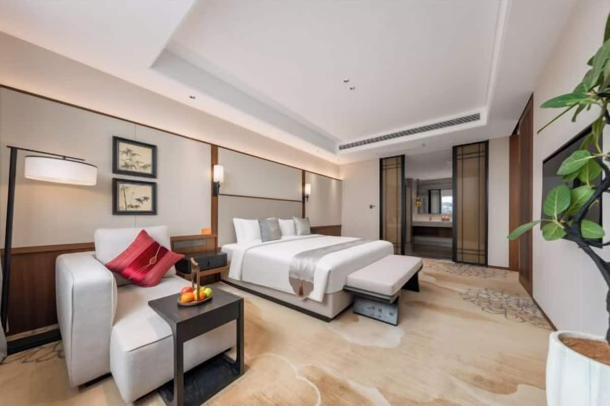 A modern hotel room with a bed, a sofa, a bedside table with fruit, a mirror, and a plant.