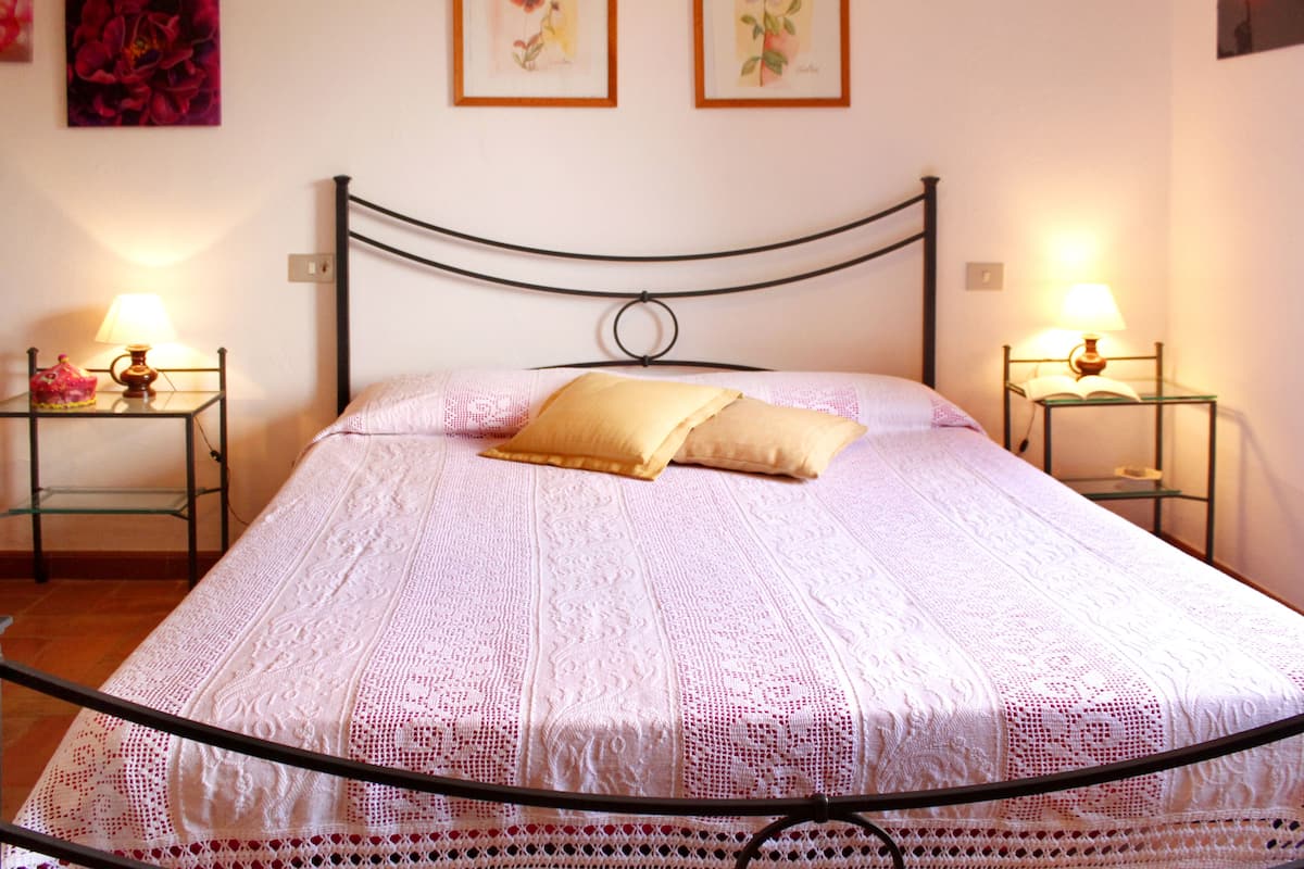 Un lit bien fait, recouvert d’une couvre-lit blanche et rose à motifs, avec deux lampes de chevet et, au-dessus du lit, une œuvre d’art encadrée représentant des fleurs.
