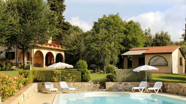 Uma área de piscina com espreguiçadeiras e guarda-sóis, cercada por vegetação e edifícios com janelas em arco.