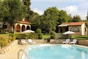 A pool area with lounge chairs and umbrellas, surrounded by greenery and buildings with arched windows.