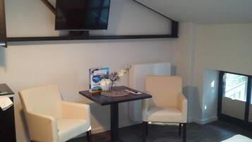 Habitación de hotel con televisor montado en la pared, dos sillones, una mesita con un florero y un folleto, y una cama con ropa blanca.