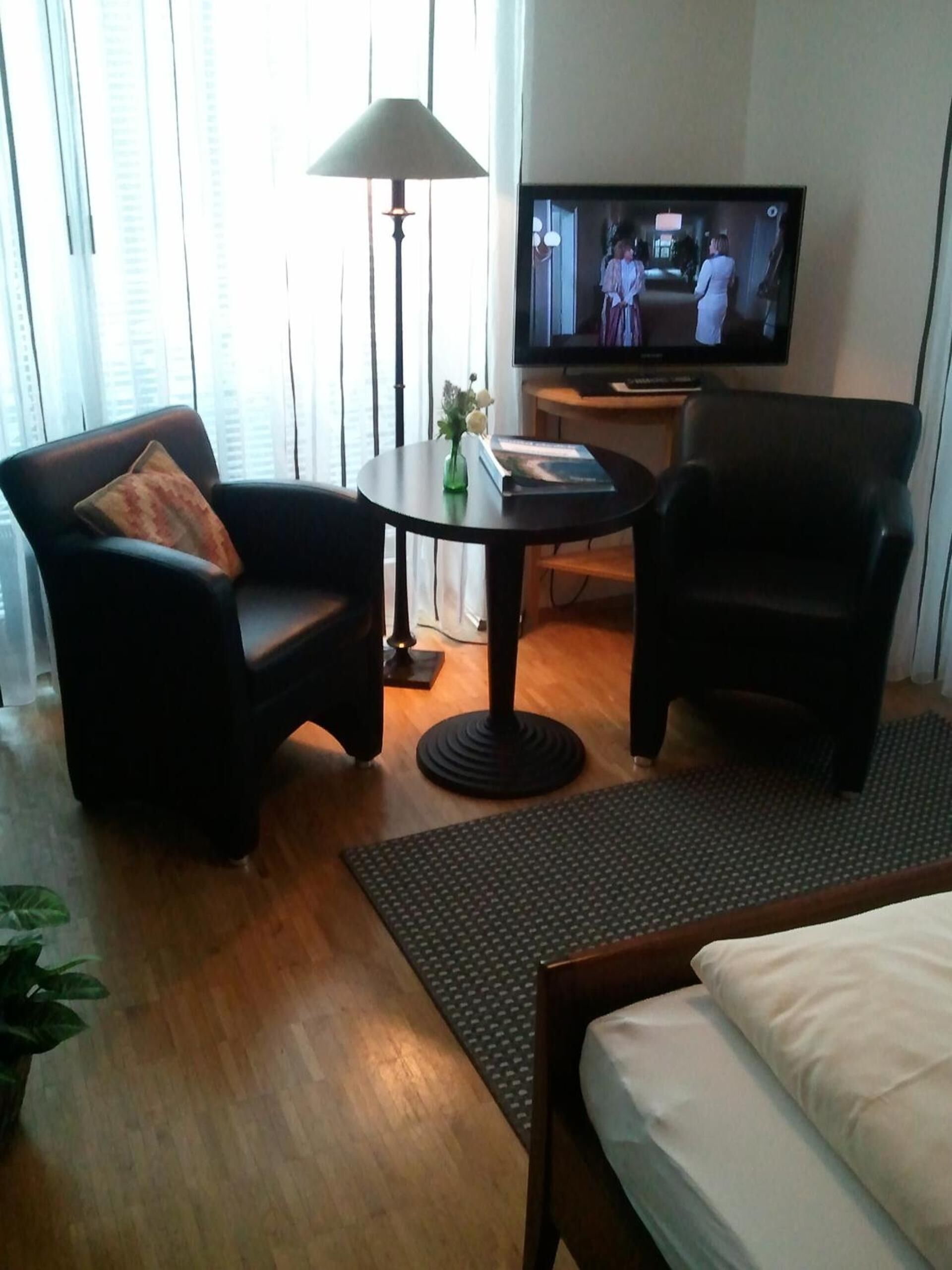 Habitación de hotel con una cama, dos sillones, una mesita con lámpara, televisor y ventana con persianas.