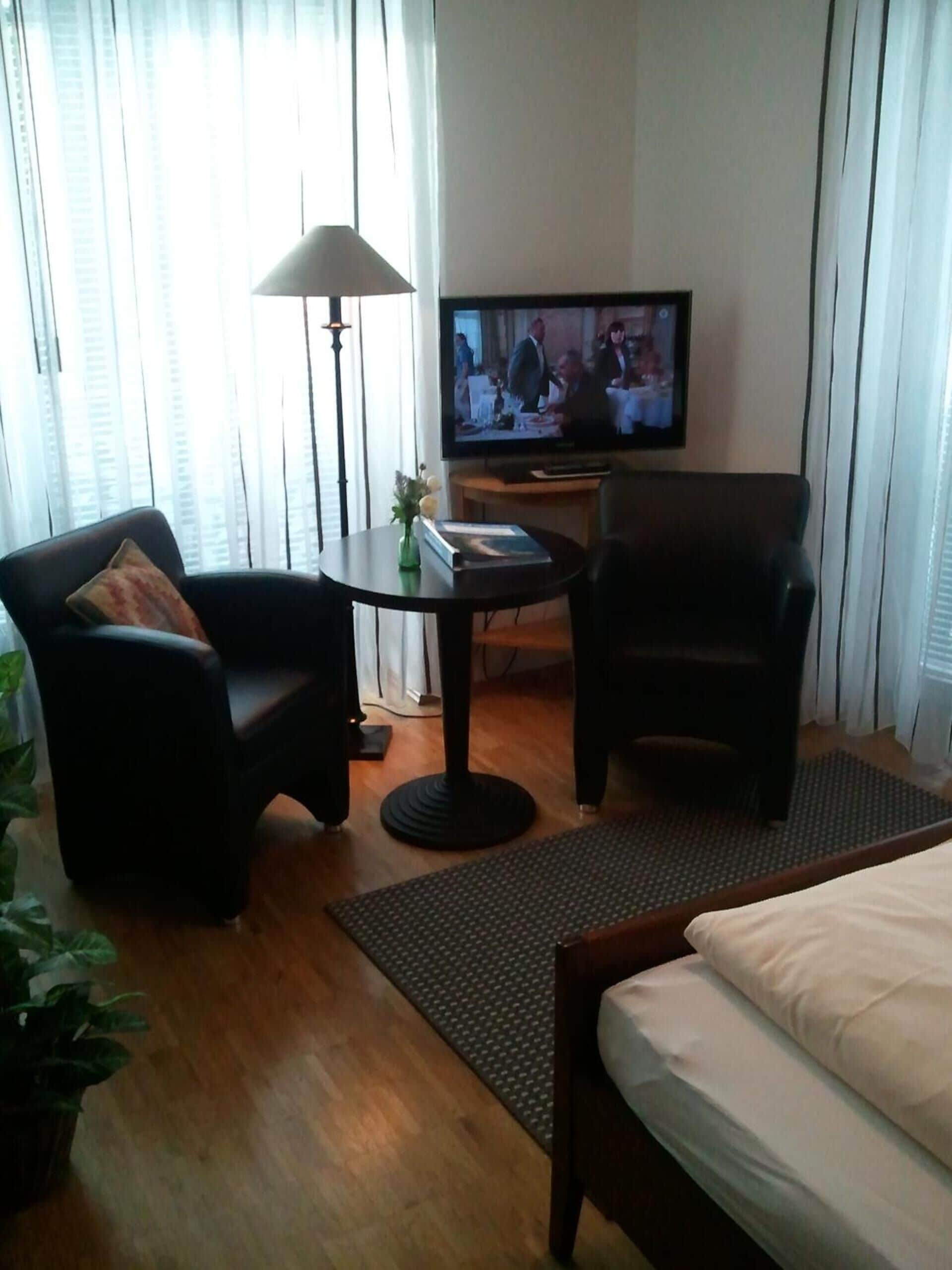 A hotel room with a bed, two armchairs, a small table with a lamp, a television, and a potted plant.