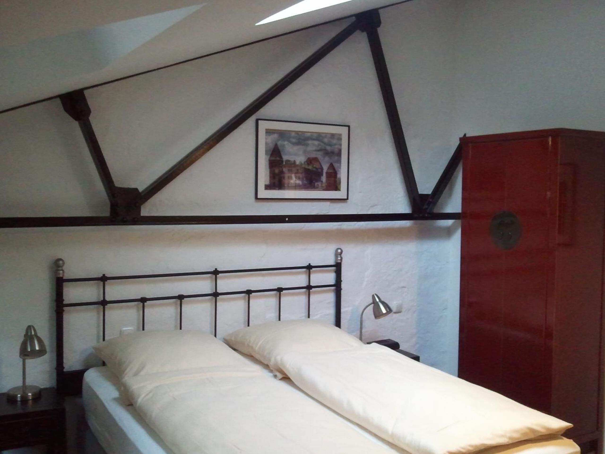 A single bed with a headboard, a bedside lamp, a nightstand, a red wardrobe, and a framed picture on the wall.