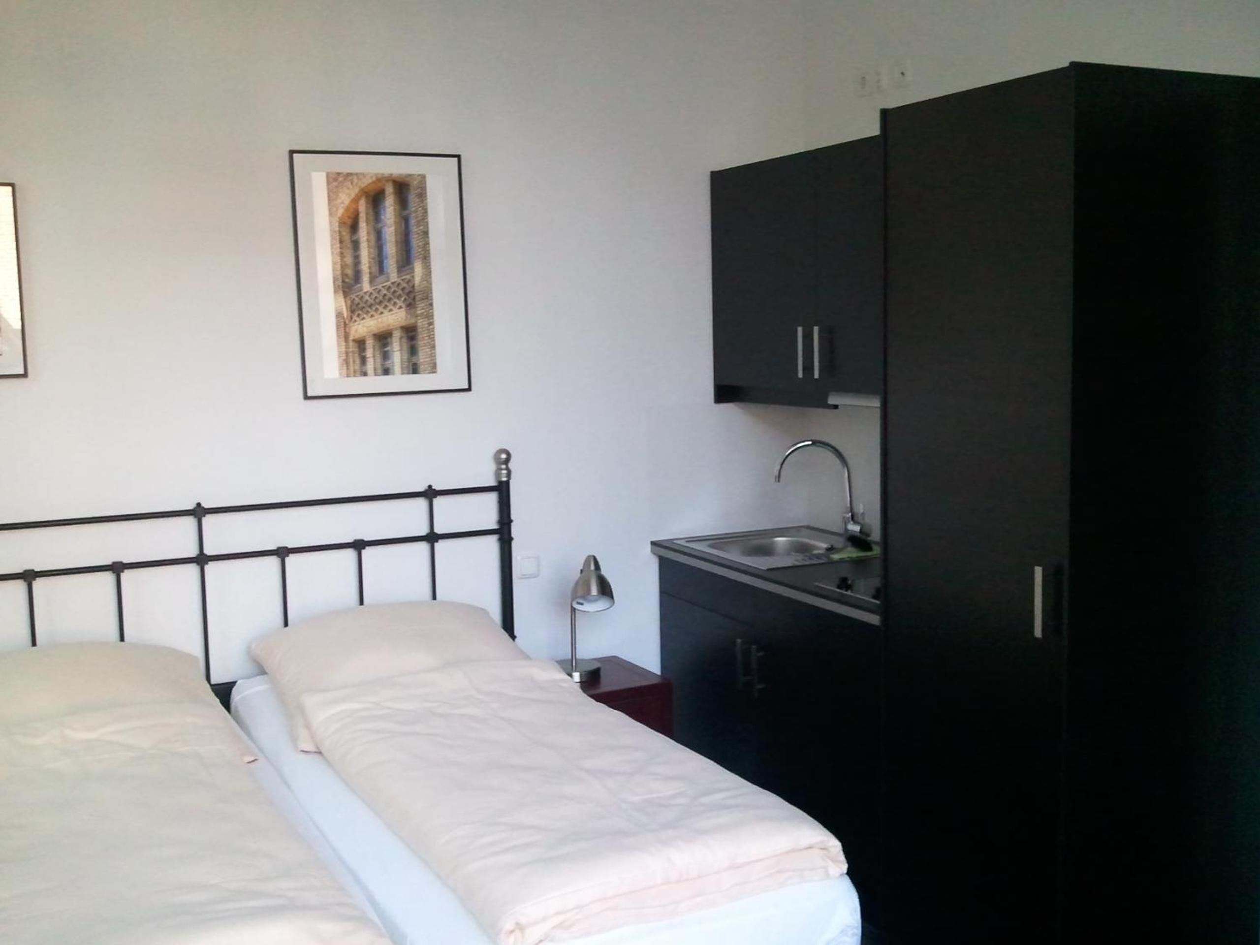 A bedroom with a bed, a nightstand, a desk with a sink, and a wardrobe.