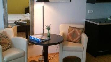 A compact living space with a round table, two armchairs, a mirror, a lamp, a framed picture, and a kitchenette.