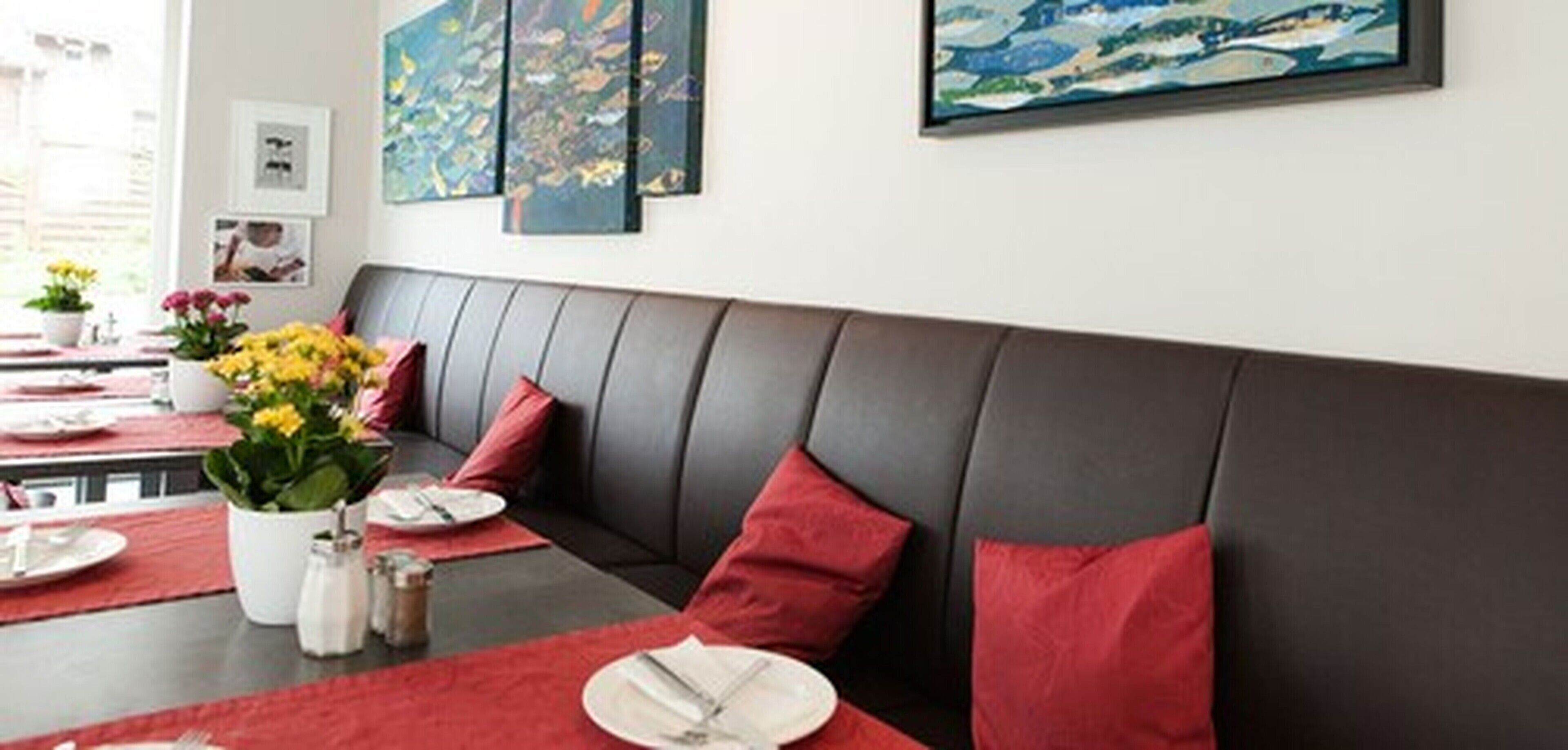 A modern dining area with a dark leather bench, red cushions, and a table set for two with plates, cutlery, and a potted plant.