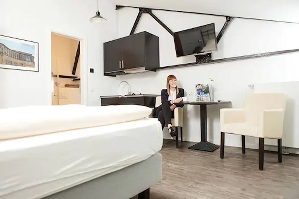 A modern hotel room with a bed, a desk, a chair, and a kitchenette.