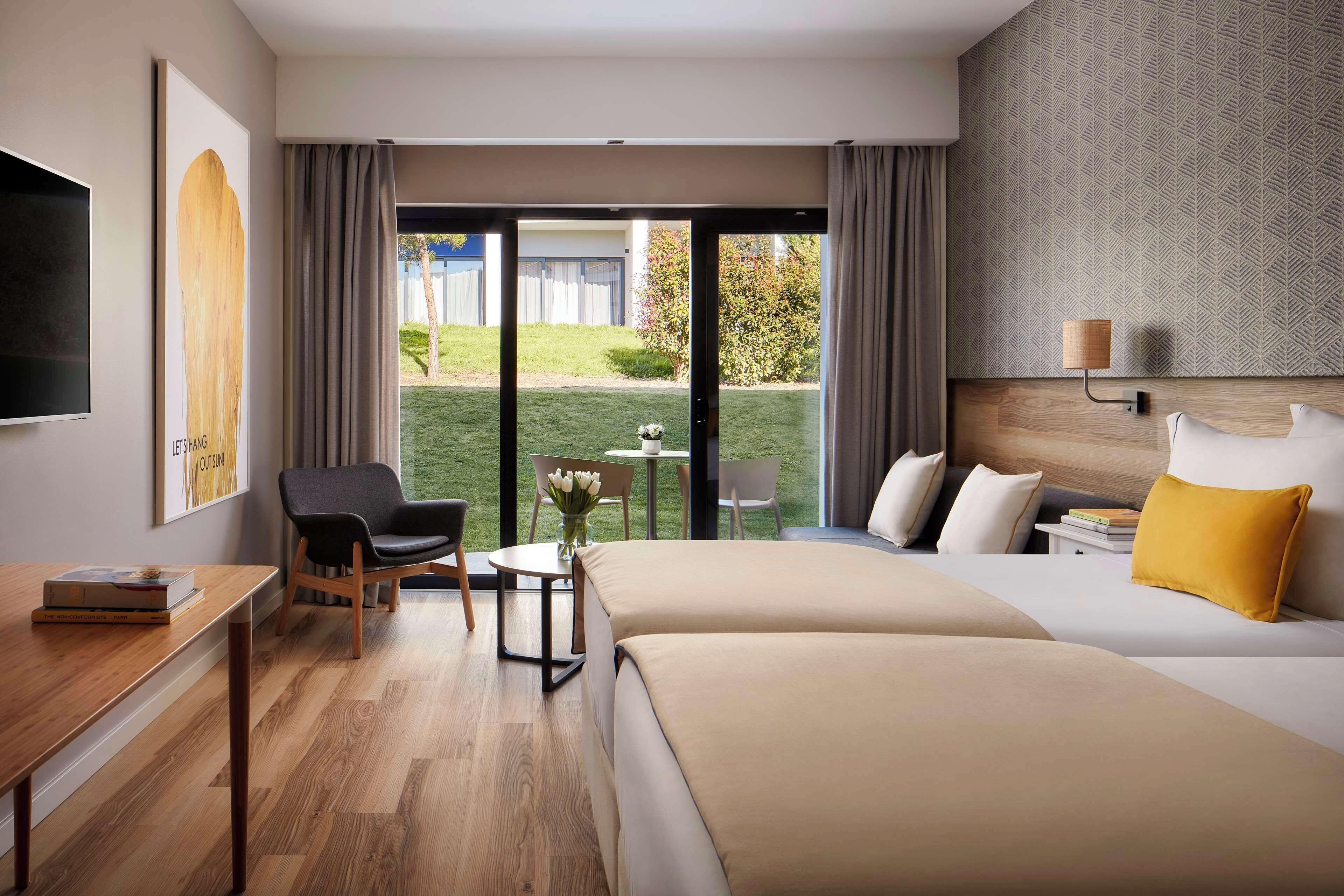 A hotel room with a bed, a desk, a chair, a small table with a vase, and a view of the outside.