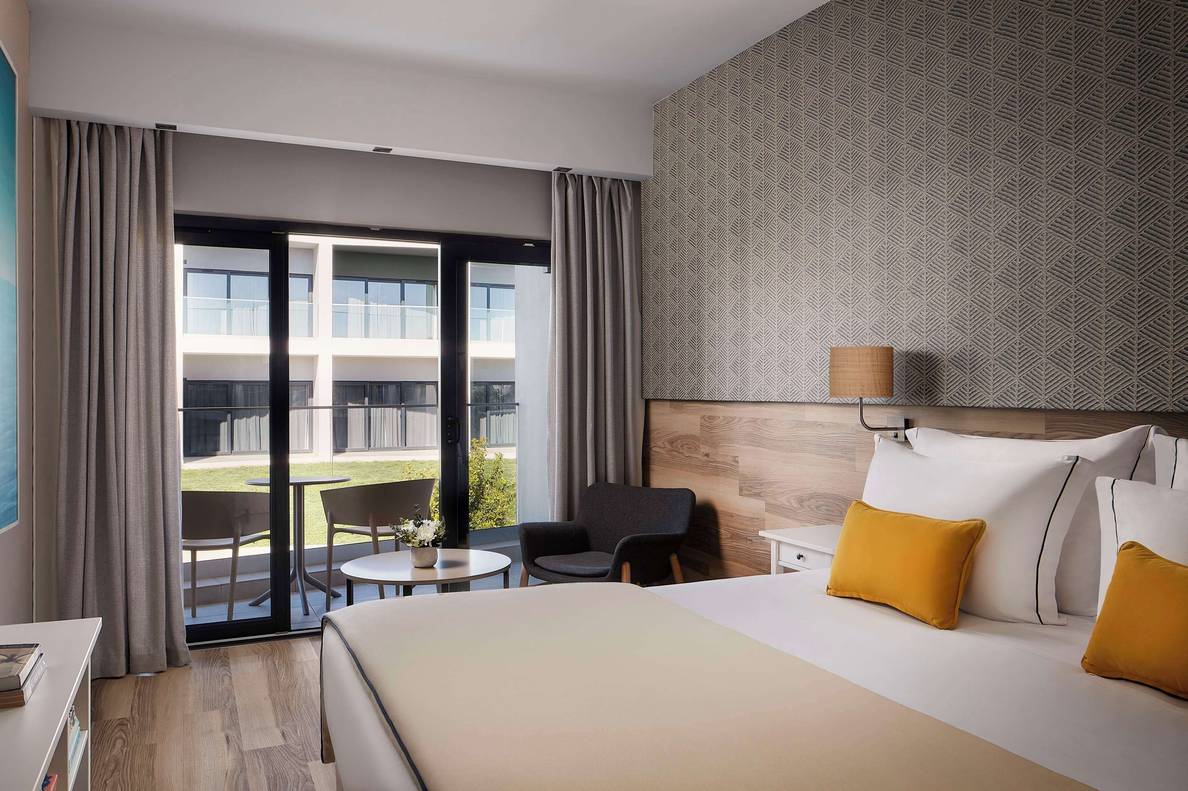 A hotel room with a bed, a desk, a chair, a small table, and a view of the outside.
