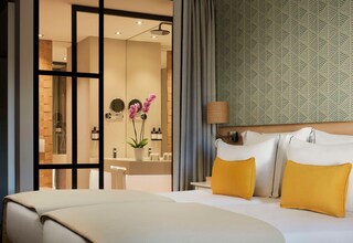 A hotel room with a bed, a nightstand, a lamp, and a bathroom visible through a glass partition.