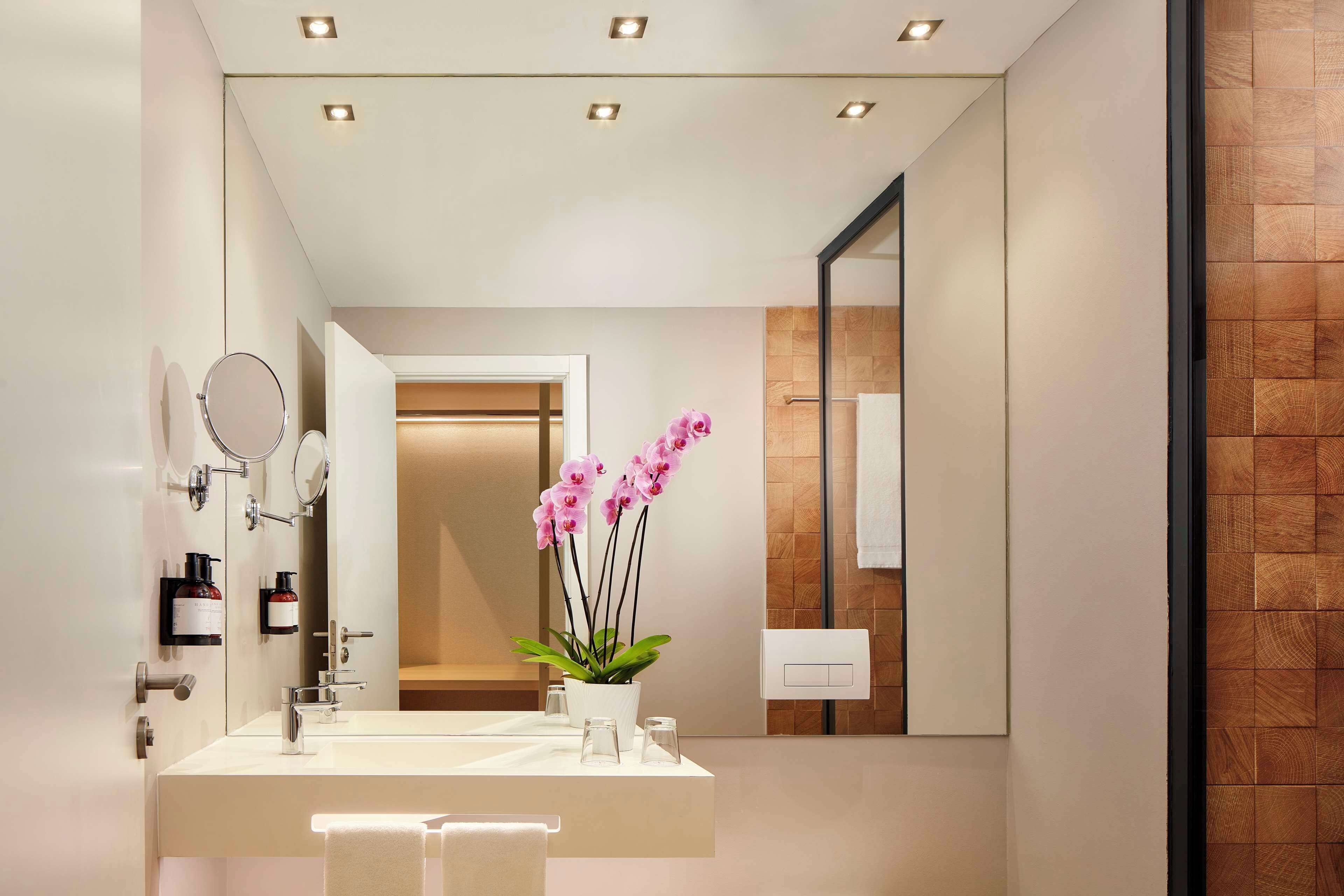 Un baño moderno con un espejo grande, dos lavamanos, una orquídea en maceta y luces empotradas en el techo.