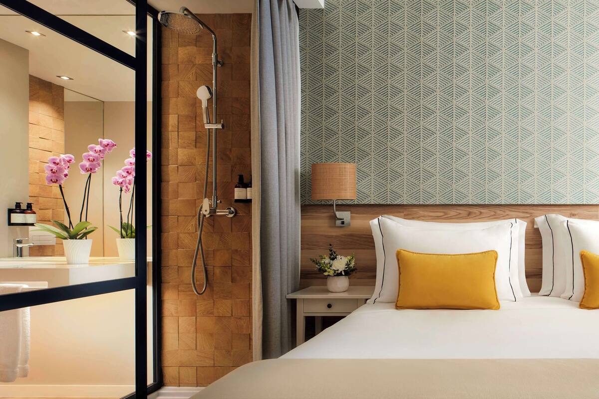 Hotelkamer met douche, bed met gele kussens, nachtkastje met lamp en een behang met patroon.