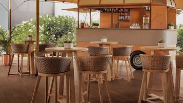Zona de bar con taburetes de madera, barra con carta de bebidas y plantas en macetas.