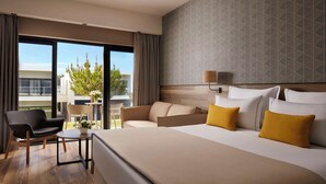 A hotel room with a large bed, a sofa, a chair, a small table, and a window with a view of greenery and buildings. - Tivoli Alvor Algarve - All Inclusive Resort (Portimão)