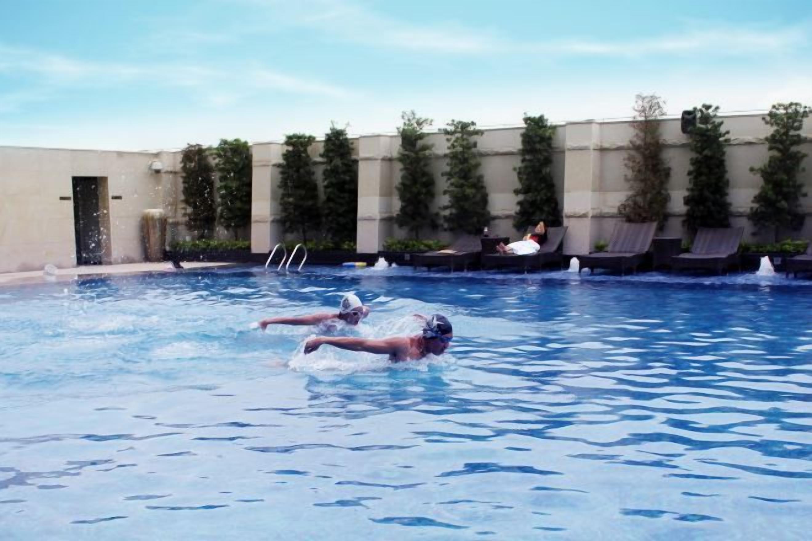 A swimming pool with two people swimming, surrounded by a building with a beige exterior and greenery.