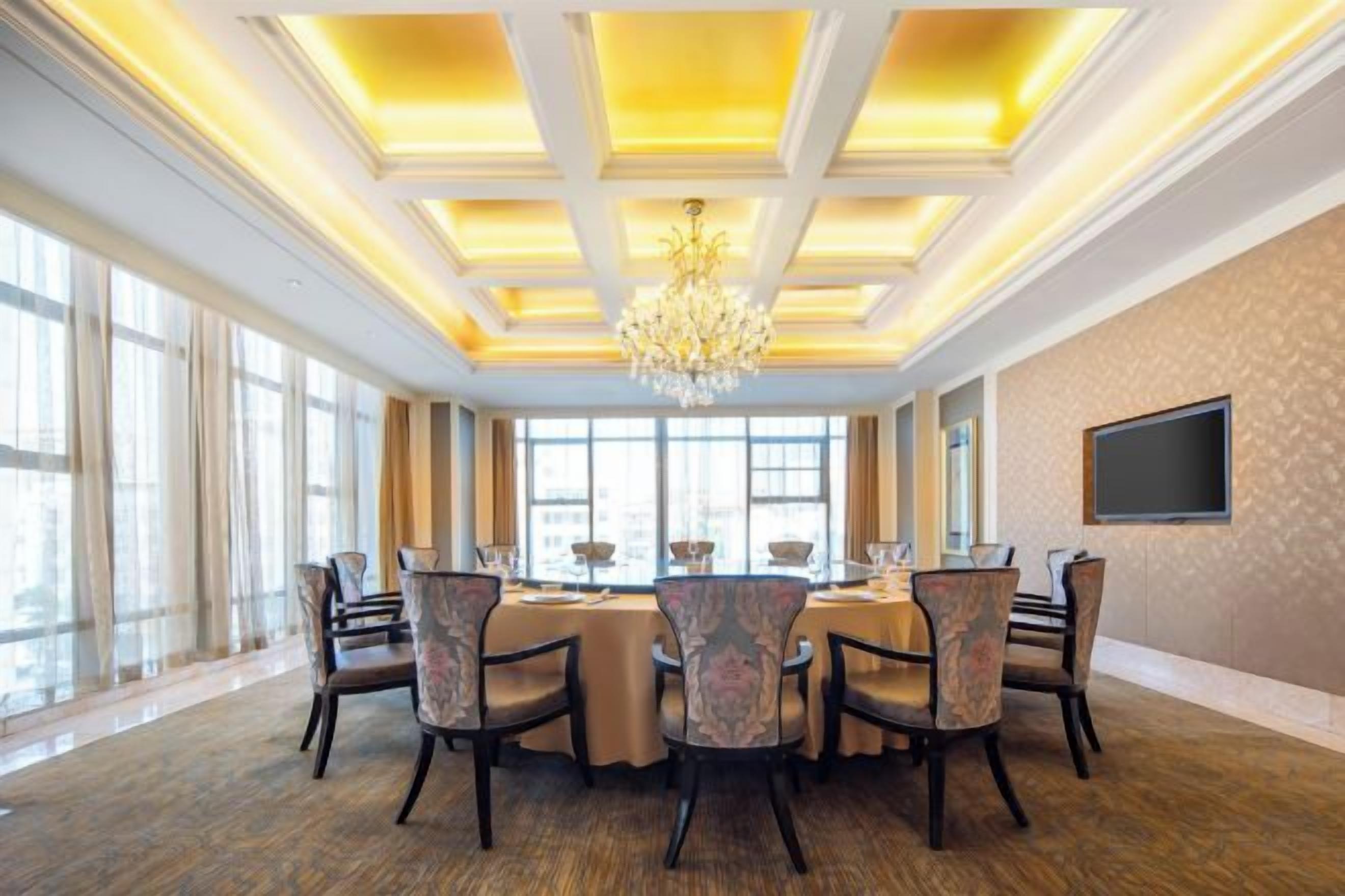 Uma sala de conferências com uma mesa grande, cadeiras, um candelabro e janelas amplas.